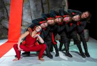 The performance piece 'Alice' presented by Jasmin Vardimon Company features a cluster of individuals positioned with their heads stacked on top of one another, tilting towards the right. The group is clad in black attire, except for the person positioned at the forefront, who wears a red outfit. Together, their bodies form a visual representation of a centipede.