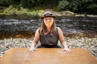 Lisette, a white woman with freckles and long brown hair, sits behind a wooden table, her hands spread out on its top. She is wearing a brown dress, a top hat and a yellow paper moustache. She is winking. The table is on a pebble beach with a river running behind it. Image credit: Rob Irish.
