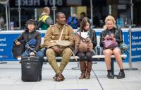 A photo of an Asian women with a suitcase, a Black service man and two white women on their phones sitting on a bench in a station