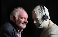 Photo of Tom Pow staring face to face with a sculpture which is wearing headphones