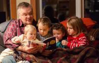 Photo shows a grandfather reading bedtime stories to three toddlers and a baby on a sofa