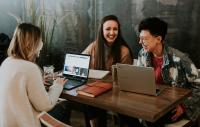 three women sitting at a table with laptops chatting and smiling