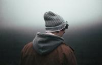 young man with woolly hat and hooded jacket facing downward and away from camera into a foggy background