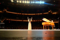 An opera singer waves to the crowd following a performance. The photo is taken from behind the singer, with a piano to their right