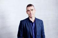 Rufus Norris, Director of the National Theatre. He is a white man wearing a blue blazer and blue shirt, with short brown hair and thick eyebrows. He is photographed against a white brick wall.