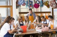 Schoolchildren and teacher sitting around table in art/design class. They are wearing blue and yellow aprons.