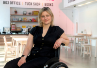 Rachel Bagshaw, incoming Artistic Director of the Unicorn Theatre, captured in the foyer. She is a wheelchair user wearing all black attire and an Apple Watch. She has pale complexion and short, blonde hair.