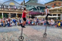 Two unicyclists surrounded by spectators juggle with clubs in front of Covent Garden Piazza