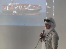 Image of woman speaking with slide of container ship in background