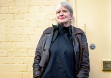 Debra King, Director at Brighter Sound. She stands against a pastel yellow wall, wearing black clothing and a leather jacket. She wears bright red lipstick and has white/grey hair.