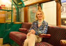 A blonde-haired woman, Elizabeth McKay, the Director and Chief Operating Officer of London Transport Museum, sits with crossed legs inside a London train exhibit. She wears a blue shirt and cream trousers.