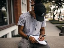 Person in balck adidas cap sitting on a bench writing in a notebook