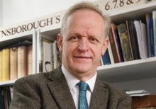 Mark Bills, incumbent Director of Gainsborough's House. He is photographed in front of a bookshelf that reads 'Gainsborough's House'. He wears a tweed, dark green/grey suit with a blue tie. He has grey hair, and is smiling at the camera.