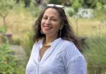Michelle Matherson, Chair of the Albany, Deptford. Matherson is stood outside in front of a a lawn; the background is blurred. Matherson has long, curly brown hair and wears a white shirt, large necklace and earrings, and sunglasses on her head. She is smiling at the camera.