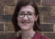 Sarah Woods, Chief Executive, Help Musicians. She has short brown hair and wears black glasses; photographed in front of a brick wall, she smiles into the camera.