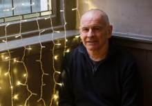 Andy Arnold, Artistic Director of Tron Theatre, sitting on a bench inside the theatre with fairy lights against a wall/window behind him. He is a bald man wearing a black jumper and is positioned to the right in the photograph.