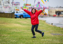 Kathryn MacDonald. A white woman pictured at a funfair. She wears a bright red coat and smiles with her arms raised.