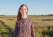 Miranda Johnson is a white woman with auburn hair. She stands in a field on a sunny day. She is smiling at the camera.