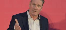 Keir Starmer speaking at a press conference. He is standing in front of a plan red background and wearing a red suit