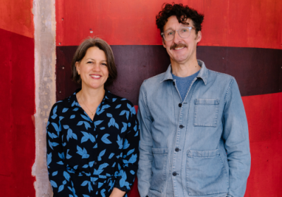 (L to R): Donmar Warehouse's Chief Executive Director Henny Finch and incoming joint Chief Executive Tim Sheader. Finch is a middle-aged white woman with dark hair. She is smiling, wearing red lipstick and a black shirt with blue birds on. Sheader is a white tall man with dark, curly hair and a moustache. He wears round grey glasses and a blue denim shirt, with a blue t-shirt underneath. They stand against a wall painted red, purple, and pink.