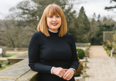 Rachel Crewes, CEO, Harewood House Trust. Crewes is a white woman with short, ginger hair and a full fringe. She wears a black dress. She is photographed outside of the House, against a wall which she is leant on.