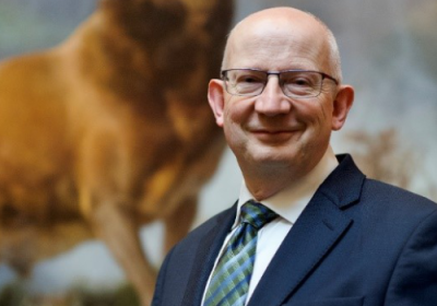 Sir John Leighton, Director-General, National Galleries of Scotland. He is a bald, white man wearing black glasses and a suit. He is standing in front of a model deer inside a museum, with a blurred background.