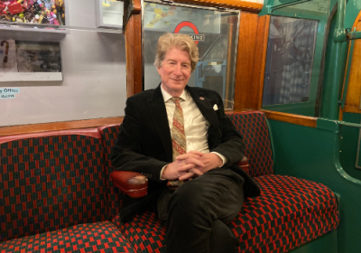 Sam Mullins OBE, CEO and Director of London Transport Museum. Mullins is a white man, photographed inside an Underground tube train replica at the London Transport Museum. He is sat down on the 'tube seat' with his legs crossed, smiling at the camera.