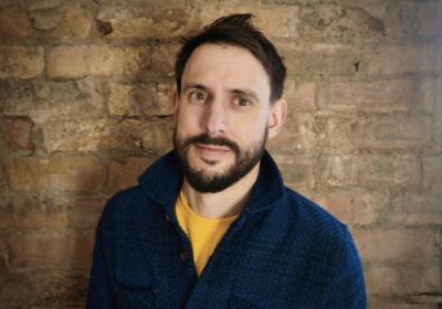 Simon Stephens has dark brown hair and a beard. He is wearing a navy cotton shirt with a yellow top underneath. He is smiling at the camera. There is a brick wall behind him.