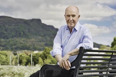 Paul Sweeney, Interim Chair, National Lottery Community Fund. He is photographed seated on a bench with a backdrop of a mountainous landscape in Northern Ireland. He is dressed in a suit.