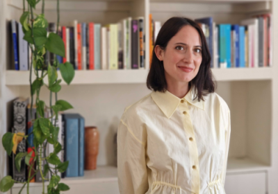Kiera Blakey, the future Director of New Contemporaries, faces the camera smiling. Her hair is short and brown, and she is wearing a white dress with a buttoned front and collar. In the background, a bookshelf and a hanging plant can be seen.
