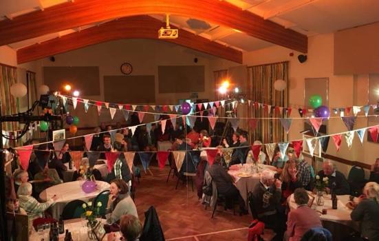 A photo of a village hall with tables of people sitting down