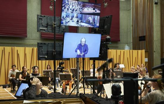 Photo of the socially distanced orchestra and screens showing a feed of the conductor