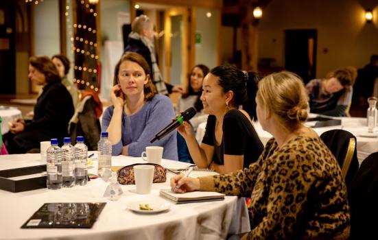 Three woman sat on a table at a work event. There are items scattered on the table including four bottles of water, mugs, a notebook, a pencil case and a small plate. Two white women are looking at an Asian woman holding a microphone (presumably speaking through it). The background is blurred, but there are other tables and guests.