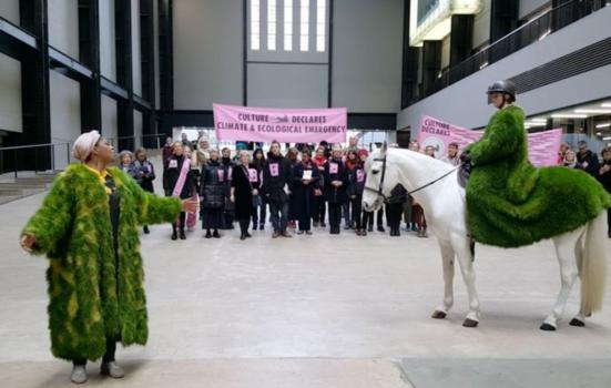 Group of people with banners in the background and two people in the foreground dress in green, leaf-like coats, one of which is on a white horse