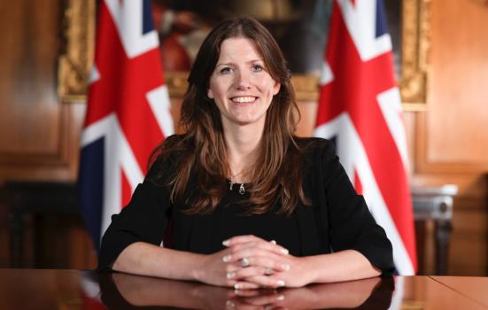 Michelle Donelan sits at a desk smiling at the camera. She sits in front of two union jacks