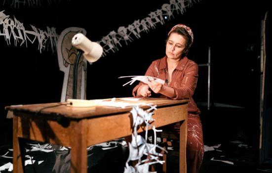 Theatre maker Viv Gordon performs Cutting Out. Viv is sat at a table cutting out images with a pair of scissors. She is wearing a brown jump suit