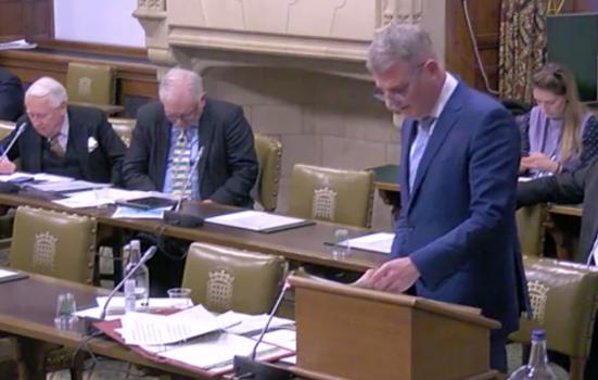 MPs listen to DCMS Minister Stuart Andrew during the debate. Andrew is standing, wearing a blue suit and reading from a sheet of paper