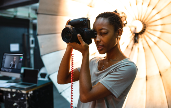 a Black woman with short black hair wearing a grey t-shirt takes a photo with an industrial camera. she is stood in front of a photographer's umbrella with a computer to her left