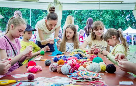Two women help a group of children with a creative project. They are working as volunteers, explaining to the children how to knit with colourful wool