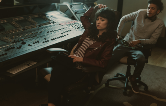 A women sits by a music recording desk, with a man sat behind her. both of them have sad expressions
