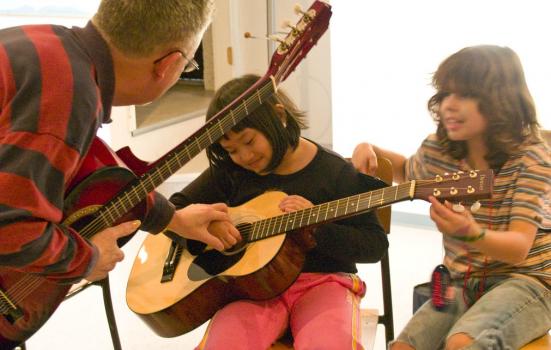 A photo of two children playing guitar next to an adult holding a guitar