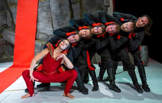 The performance piece 'Alice' presented by Jasmin Vardimon Company features a cluster of individuals positioned with their heads stacked on top of one another, tilting towards the right. The group is clad in black attire, except for the person positioned at the forefront, who wears a red outfit. Together, their bodies form a visual representation of a centipede.