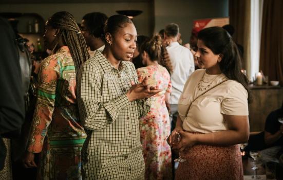 A Black woman speaking to a mixed-race woman at a social event. The Black woman is wearing a tartan dress with her hair in a tight bun, while the mixed-race woman wears a white top & skirt and has her hair down.