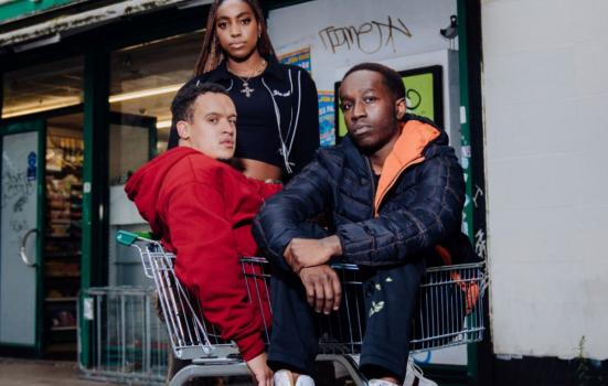 National Youth Theatre's production shot from 'GONE TOO FAR!'. There are two young men sitting in a trolley and a young women standing behind; they are all looking into the camera. Corner shop in the background.