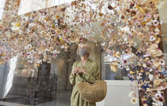 A woman in a floral dress and face mask looking an art installing of floating flowers