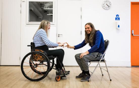 Image shows two people seated, one in a wheelchair, touching hands