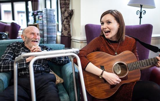 a woman seated playing guitar to an elderly gentleman seated witha zimmer frame in the foreground