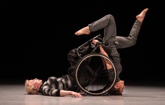 A female wheelchair-using dancer rests on her back, another female dancer balances on her head using the wheelchair for support
