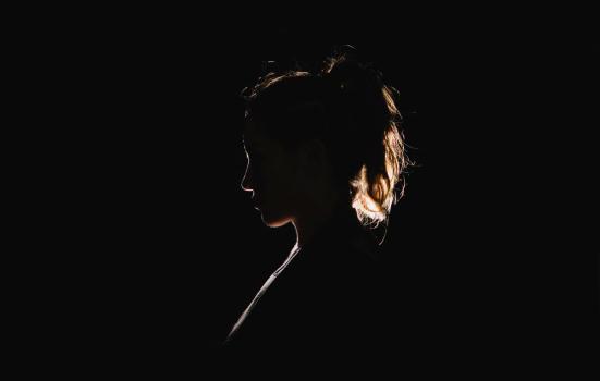 The contour of an individual who appears to be a woman, standing in the darkness with a spotlight positioned behind her, creating a shadow that outlines her figure like a silhouette.