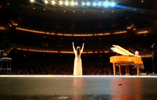 An opera singer waves to the crowd following a performance. The photo is taken from behind the singer, with a piano to their right
